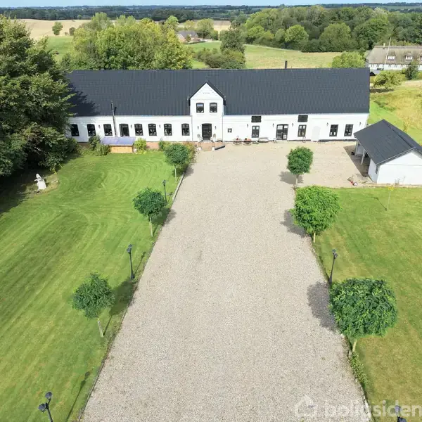 Et hvidt, langstrakt landhus med sort tag står stille i en have med grønne plæner og træer. Foran huset er en grusbelagt gårdsplads.