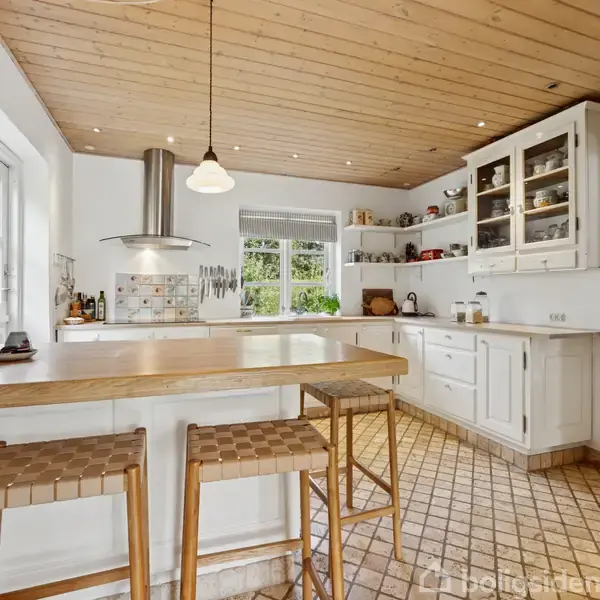 Køkken med træloft og flisegulv har en køkkenø med høje stole, overskabe med glaslåger og et vindue med udsigt. En plakat med "MORRIS MINOR" hænger på væggen.