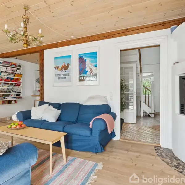 Blå sofaer står i stue med træloft og trægulv. Pejs brænder ved siden af sofabord med frugtskål. Vægdekorationer inkluderer plakater og magasinhylder. Dør fører til gang.