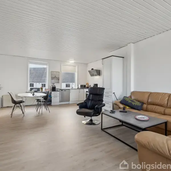 En stue med en brun lædersofa og sort lænestol omkring et sofabord. I baggrunden ses et spisebord ved vinduet, og et åbent køkken. Lys træfarvet gulv og hvide vægge.