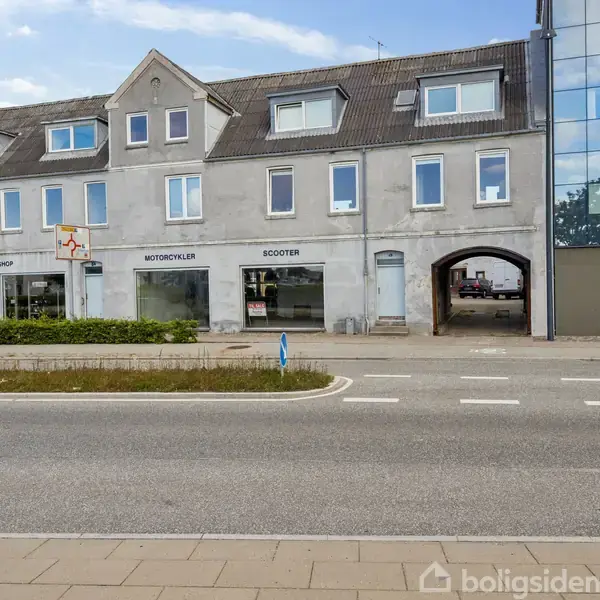 En grå bygning med butiksvinduer, inklusive en motorcykelforretning, står ved en vej med fortov. En buegang fører ind til en baggård.