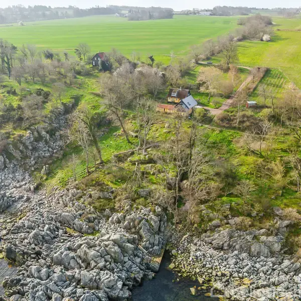 Et hus ligger fredeligt ved siden af klippeformationer langs kysten. Omgivet af grønne marker og træer, skaber det en harmonisk balance mellem natur og landlig idyl.