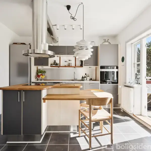 Køkken med moderne grå skabe, træbord og en emhætte, beliggende i et lyst rum med store vinduer. To stole står ved køkkenøen, og der hænger en kunstnerisk lampe over bordet.