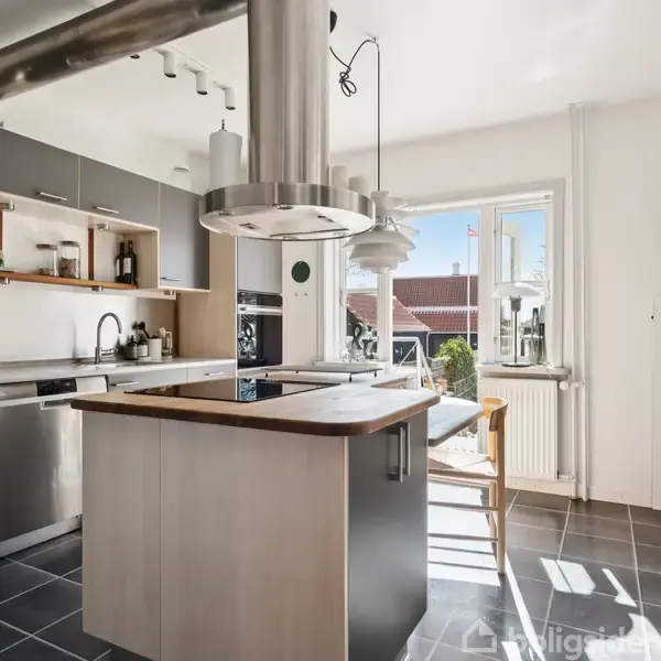 Køkkenø, moderne stil, står centralt med emhætte over. Omkring er mørkegrå skabe, stålopvaskemaskine, og hvid væg med stort vindue, der tillader naturligt lysindfald.