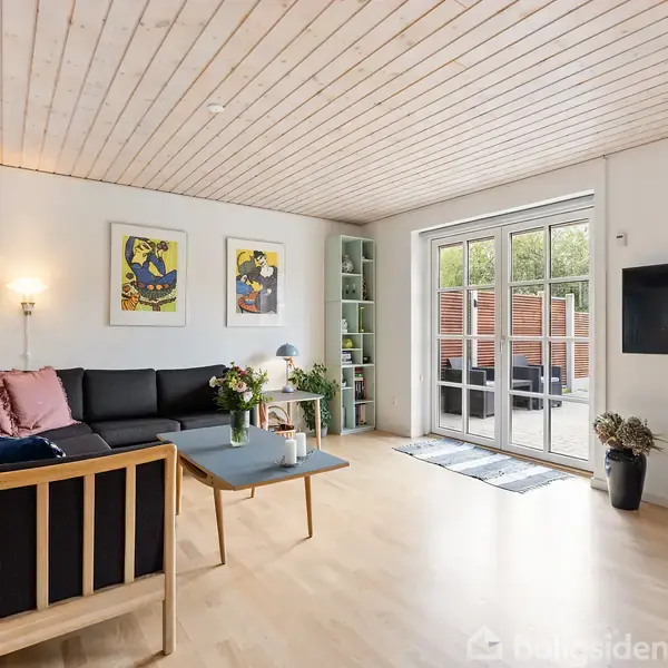 Sort sofa står i hjørnet med farverige puder og et sofabord foran. Vægdekoration og hylde med planter i et lyst rum med trægulv og stor glasdør.