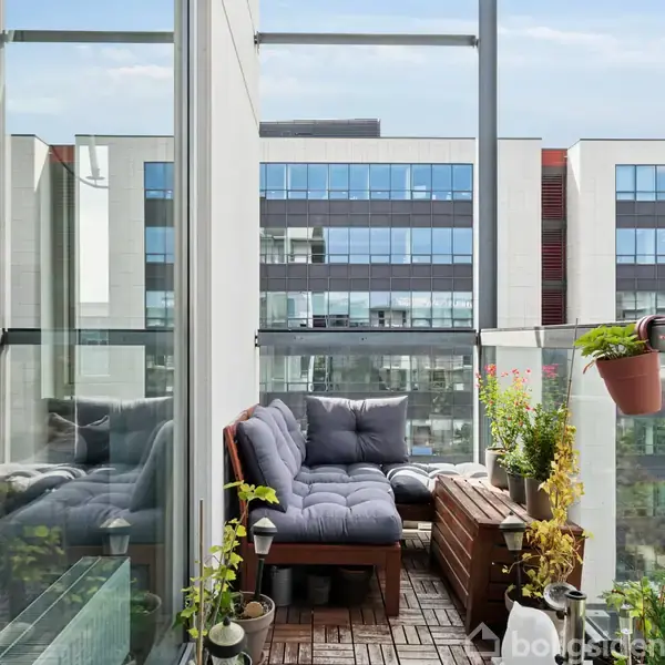 Sofa med grå puder står på en balkon, omgivet af glasrækværk og planter i krukker. Moderne bygninger med store vinduespartier ses i baggrunden.
