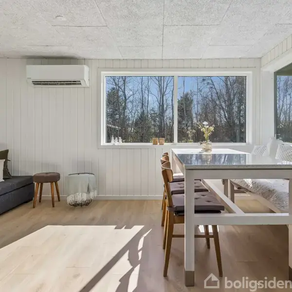 Stue med hvidmalede vægge og store vinduer. Sofa står ved venstre væg, spisebord med stole ved vinduet. Udsigt til træer udenfor. Lys træparket på gulvet.