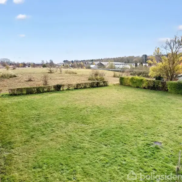 Græsplæne breder sig ud, omgivet af hække og træer, med en åben mark og flere bygninger i det fjerne under en blå himmel med få skyer.