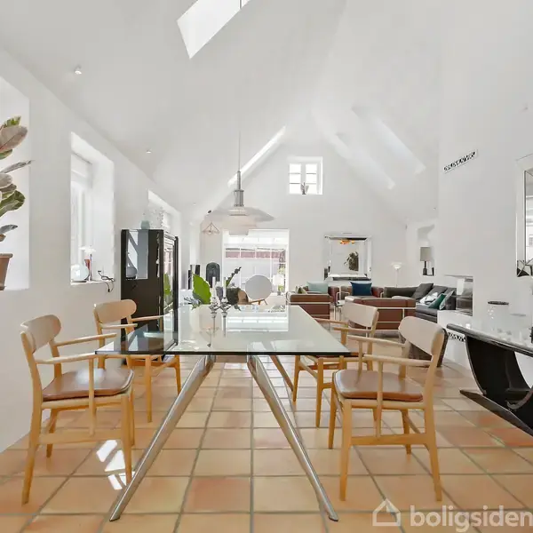 Et moderne spisestuemiljø med glasbord omgivet af træstole. Rummet er lyst med skråloft og store vinduer. Planter og kunstgenstande pryder omgivelserne i det rummelige opholdsområde.