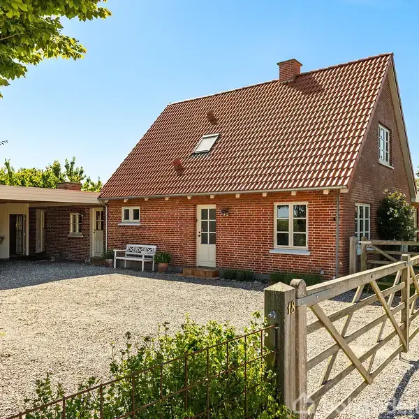 Rødt murstenshus med tag af røde teglsten står stille. Gruset indkørsel omgivet af grønne træer. En garage med hvid port til venstre for huset.