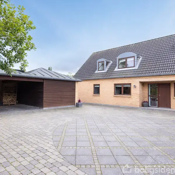 Et enfamiliehus med røde mursten og sort tag ligger ved siden af en dobbelt carport. Foran er en stor, flisebelagt gårdsplads med en hvid bil parkeret.