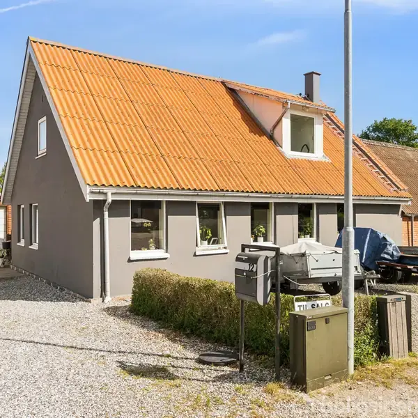 Et gråt hus med rødt tegltag står i solskin. Foran huset er der grus på gårdspladsen, en lille busk og en postkasse. Til venstre ses en garage.