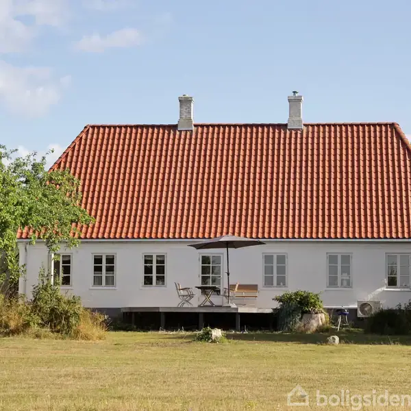 Hvid hus med rødt tegltag, står i en grøn have med træer. Terrasse med havemøbler ved huset, og græsplænen omgiver det.