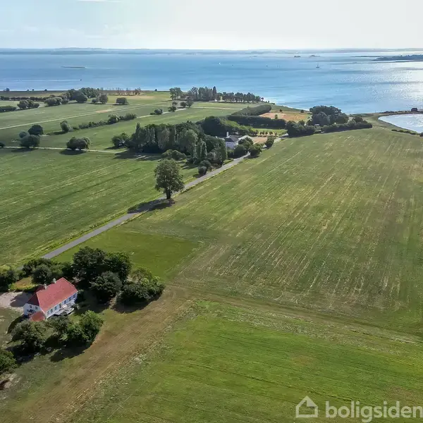 Hus med rødt tag ligger i et grønt landskab. Omkringliggende marker strækker sig mod fjerne træer og en kystlinje med udsigt over vandet under en blå himmel.