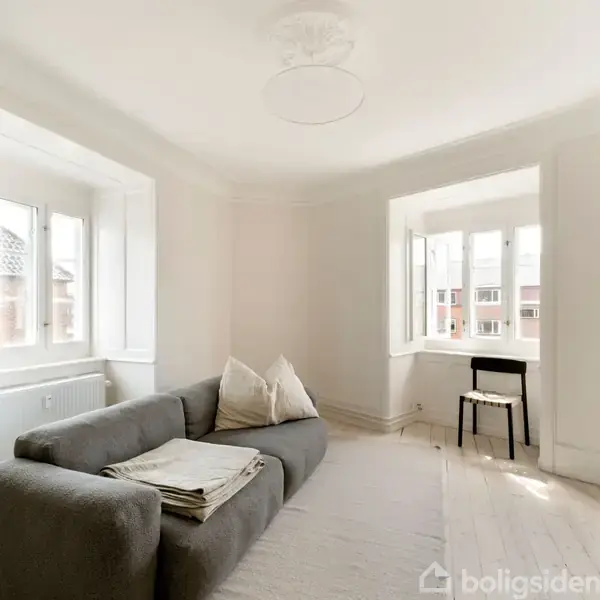 Grå sofa med puder står i et lyst rum med to store vinduer. Der er en stol og et lille bord ved det ene vindue og en reol med bøger.
