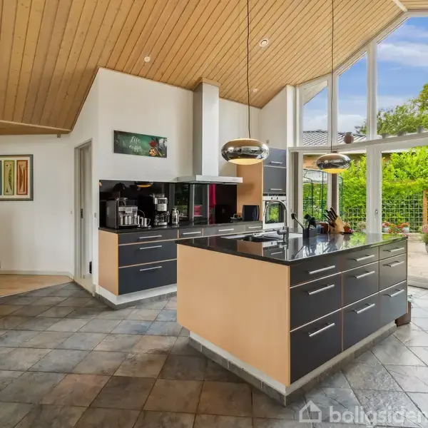 Et køkken med en central sort køkkenø, moderne apparater og hængende lamper. Store vinduer med udsigt til en grøn have og skrå loftpaneler tilføjer lys og rummelighed.