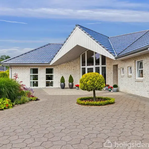 Hvid murstenshus med blå tagsten står på en flisebelagt gårdsplads. En rund, grøn busk er centralt placeret, omgivet af blomsterbede under en klar blå himmel.