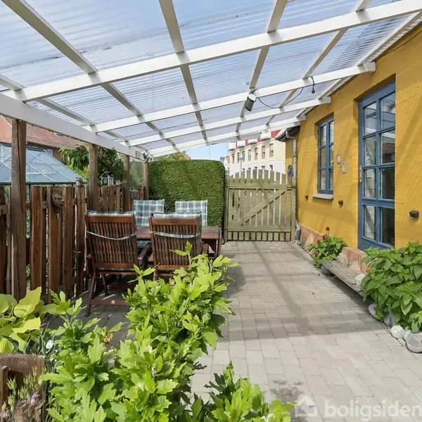En overdækket terrasse med borde og stole under et transparent tag. Til venstre et hegn og et drivhus, til højre en gul bygning med blå vinduer og planter ved siden.
