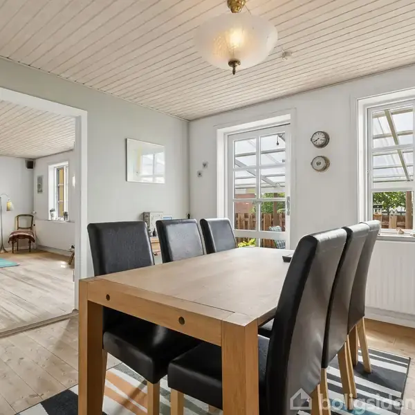Spisebord med sorte stole står i et lyst rum med trægulv. Store vinduer og glasdør fører ud til en overdækket terrasse. Stue med sofa i baggrunden.