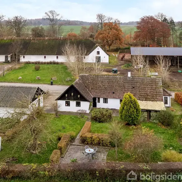 Et bindingsværkshus med stråtækt tag står i en frodig have omkranset af hække og træer. Baggrunden har landsbyhuse og en åben mark i efterårsfarver.