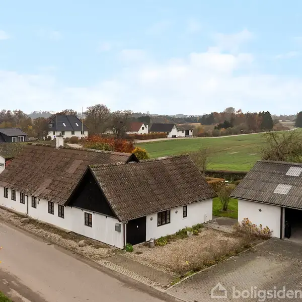 Et hvidt bindingsværkshus med sort bindingsværk står ved en landevej. Til højre ses en åben garage. Omgivet af marker og spredte træer under en klar himmel.