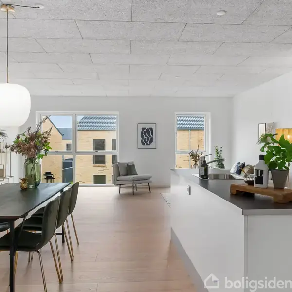 Spisestue i moderne design med sort bord, seks stole og hængelampe. Åben forbindelse til lyst køkken med planter. Bagved ses store vinduer der viser bygninger udenfor.