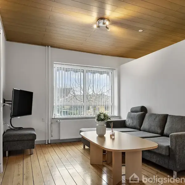 Gråt hjørnesofa står i en stue med lys trægulv og træloft; et rundt sofabord står foran sofaen. Vindue med udsigt dominerer rummet, ved siden af hænger et vægmonteret TV.