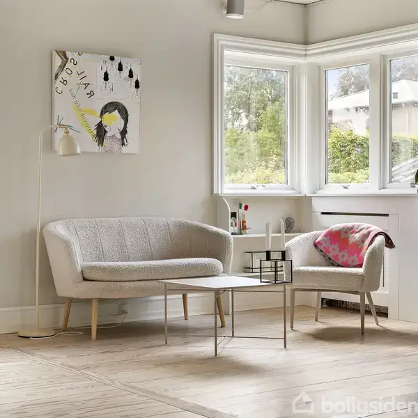 To stole og et bord står i en lys stue med kunst på væggen. Store vinduer giver udsigt til en grøn have.