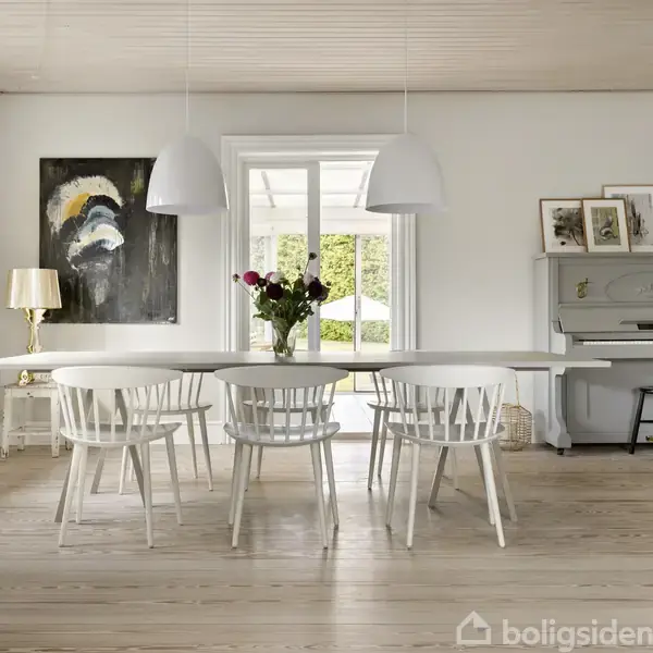 Et lyst og luftigt spiseområde med et hvidt bord omgivet af seks hvide stole. To hængelamper lyser rummet op, mens en vase med blomster pryder bordet. I baggrunden ses et klaver og kunstværk.
