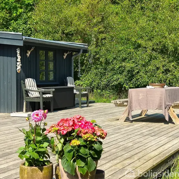 Terrasse med træmøbler står udenfor et lille, mørkt bjælkehus omgivet af frodig vegetation. Bordet er dækket med dug, og blomster i krukker pryder området.