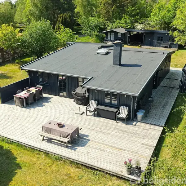 Sort sommerhus med skorsten står på en rummelig træterrasse, omgivet af frodig have og træer. Terrasse har udendørs møbler og grill, hvilket giver en hyggelig atmosfære i naturen.