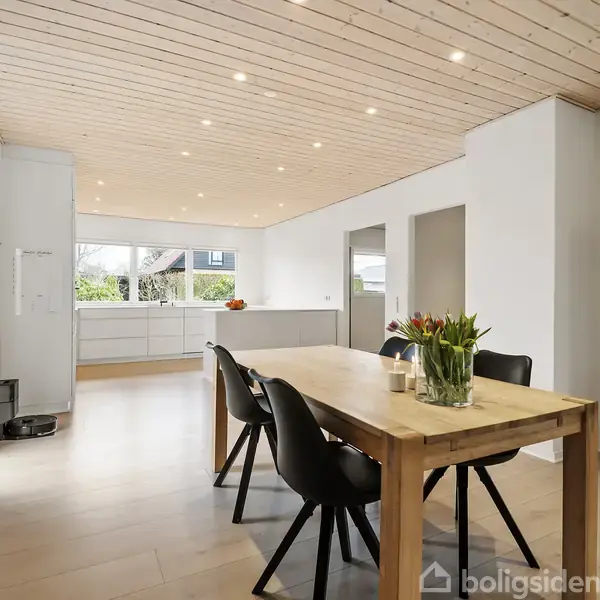 Et spisebord med sorte stole står i et lyst køkken-alrum. En vase med tulipaner pryder bordet. Et moderne, hvidt køkken ses i baggrunden med store vinduer og en træloft.