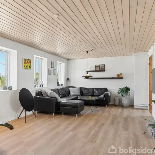 En moderne stue med en sort lædersofa i hjørnet, omgivet af hvide vægge og træloft. Der er flere kunstværker, en gulvlampe og planter i rummet.