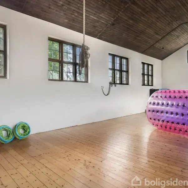 En stor, tom gymnastiksal med trægulv, hvor en farverig, oppustelig cylinder ligger. Ringe hænger på en væg. Vinduer giver naturligt lys. En basketballkurv ses i baggrunden.