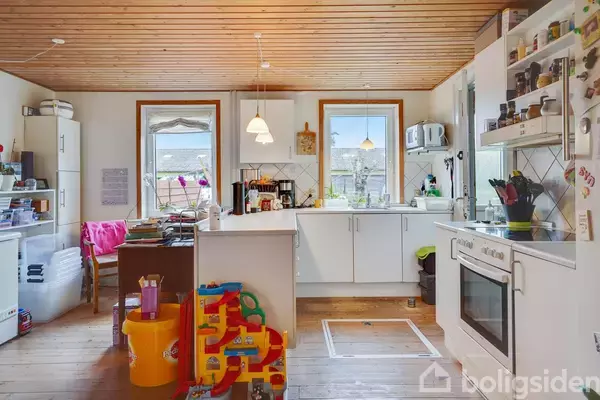 Køkken med hvide skabe og træloft, legetøj samlet på gulvet. To vinduer slipper lys ind, med udsigt til udenfor. Diverse køkkenredskaber og apparater er på hylder og borde.