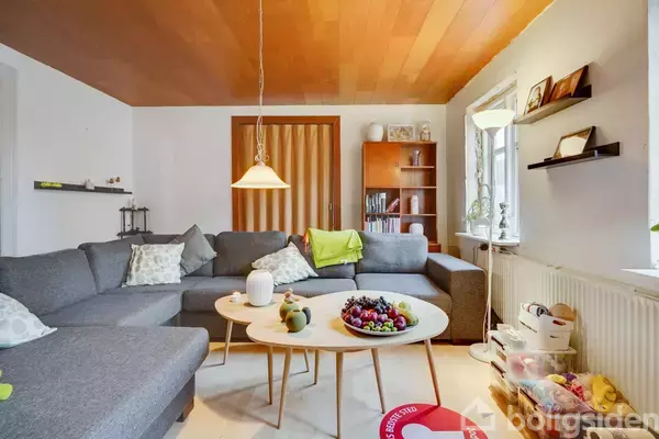 Stue med grå hjørnesofa, sofaborde pyntet med frugter, omgivet af reoler, lamper og hylder. Loftet er i træ, og store vinduer giver lysindfald fra vindueskarmen med dekorationer.