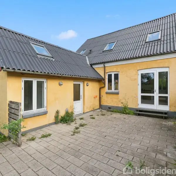 Et gulmalet hus med grått tag står stille på en brolagt gårdsplads. Døre og vinduer er hvide, omgivet af lidt vildtvokset vegetation og blå himmel.