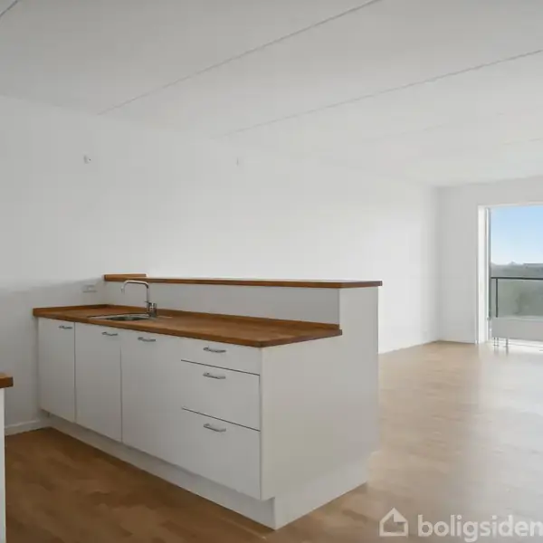 Et moderne køkken med hvide skabe og træbordplader er inaktivt ved siden af en stue. Stuen har en lys atmosfære med en stor altandør, der åbner ud til en udsigt over byen.