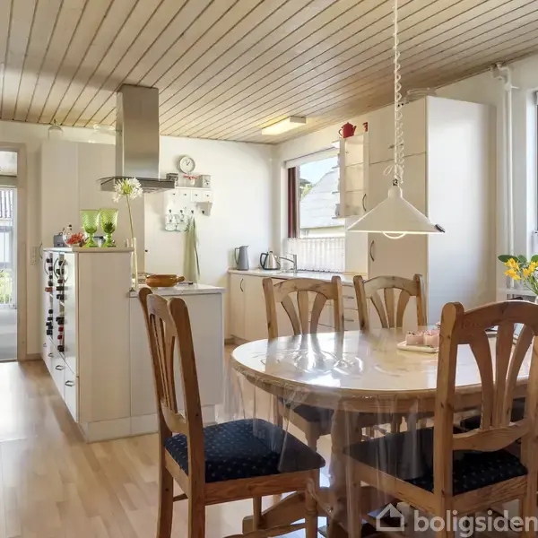 Rundt spisebord med seks stole er placeret i et lyst køkkenalrum. Træloft og lyse trægulve skaber en hyggelig atmosfære. Vinduerne giver udsigt til en solrig have.