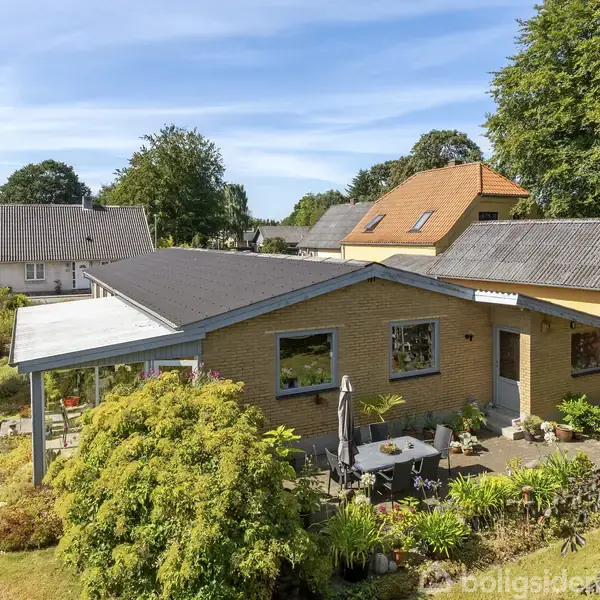 Et enfamiliehus med gult murværk og sort tag ses i en frodig have med terrassemøbler. Omgivet af andre huse på en rolig villavej med træer i baggrunden.