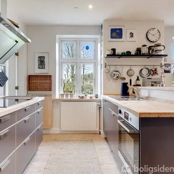Et moderne køkken med grå skabe og træbordplader. Køkkenet er lyst med store vinduer og hylder med køkkengrej. En ur hænger på væggen. Planter og billeder dekorerer rummet.