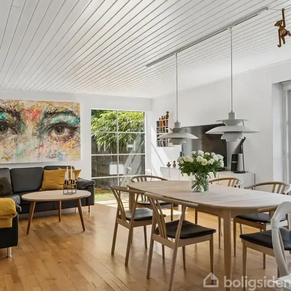 Stue med hvidt loft og trægulv, hvor et spisebord er omgivet af stole. Sofa med maleri af øjne hænger på væggen. Store vinduer slipper lys ind fra haven.