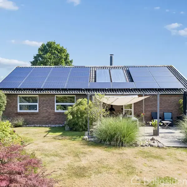 En enetages murstenshus med solpaneler på taget. Træterrasse med solsejl, omgivet af en velplejet have med grønne buske og fritstående stole. Klar blå himmel i baggrunden.