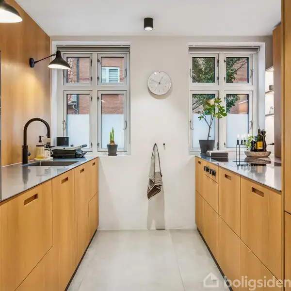 Køkken med træskabe, sort vandhane og lys bordplade. Handling: Ingen åbenlys handling. Kontekst: To vinduer med planter og vægur på væggen, lyse gulvfliser.