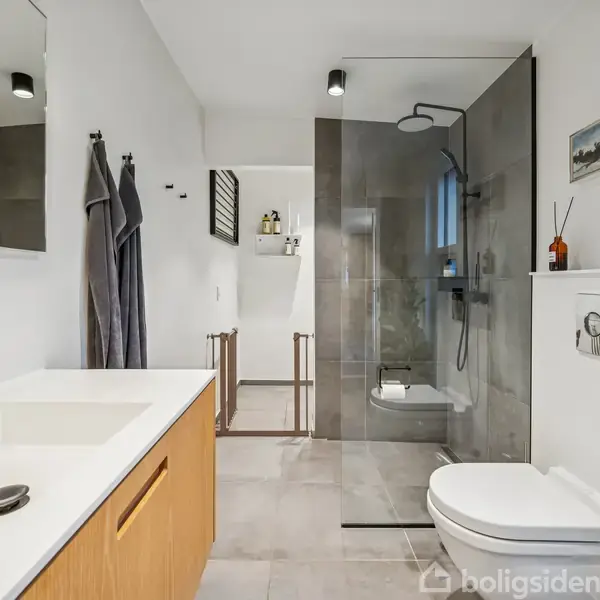 Badeværelse med vask og sort armatur til venstre; bruser i glasindkapslet område i baggrunden; håndklæder hænger på væggen; toilet til højre; dekorativ hylde og maleri over.