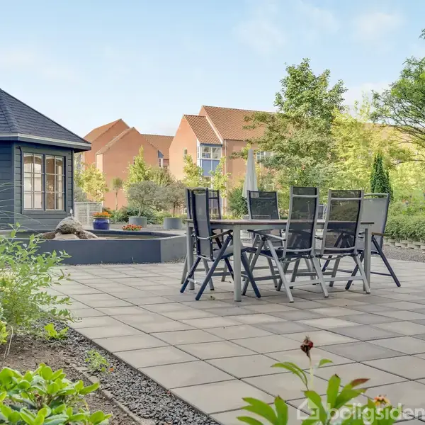 Pavestole og bord står på en flisebelagt terrasse, omgivet af frodig have med græs og buske, mens en lille pavillon står i baggrunden under en blå himmel.