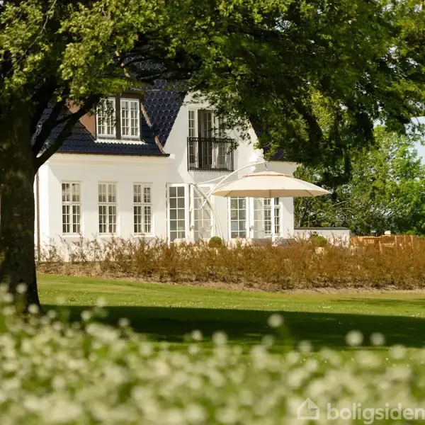 Hvidt hus med sort tag står stille omgivet af træer og frodigt grønt græs. En terrasse med parasol ses i forgrunden, hvilket skaber en rolig atmosfære.
