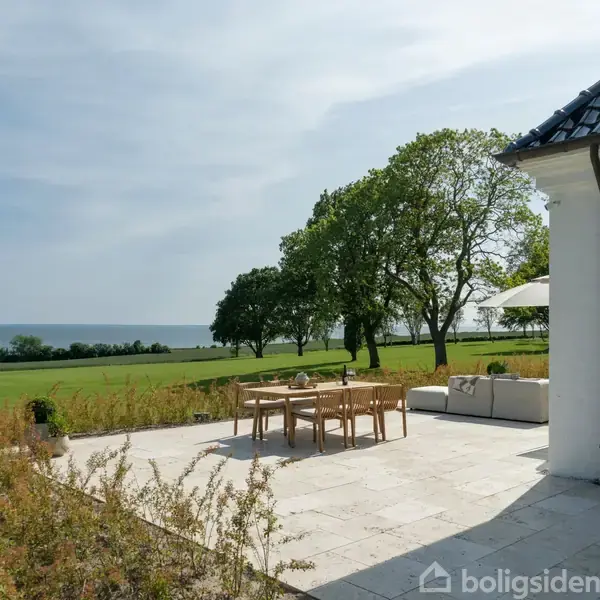 Terrassen har et spisebord med stole og parasol. En sofa står ved siden af. Den ligger ved et hus ud mod en stor grøn græsplæne med træer, havet i baggrunden.