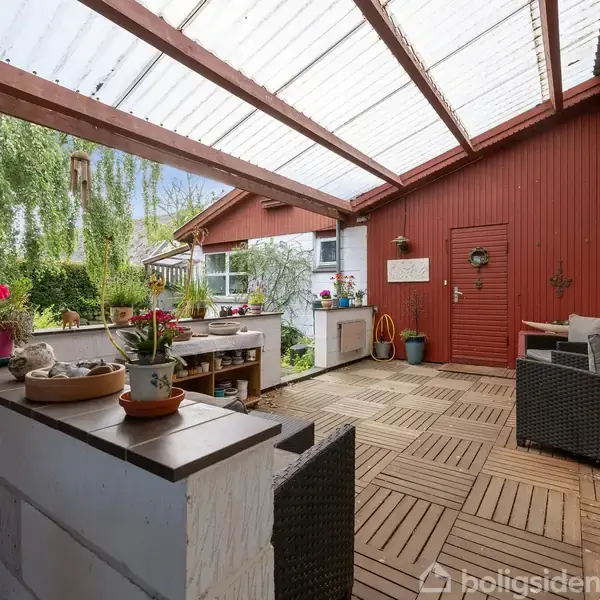 Overdækket terrasse med gennemsigtigt tag; blomster og planter på træbænk og køkkenbordlignende område. Rød trævæg med dør i baggrund; fletmøbler til afslapning.