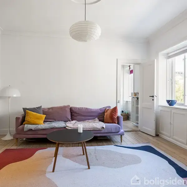 En stue med en lys, moderne sofa fyldt med farverige puder står på et trægulv. Et rundt sofabord er foran sofaen, omgivet af et stort, mønstret tæppe. Vinduerne lader lys strømme ind.